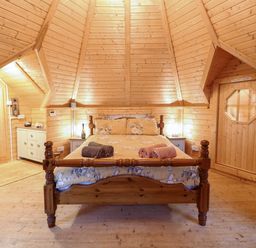 A bedroom with a wooden bed, rolled towels on the bed, wooden walls and ceiling, a cushioned chair and storage cabinets at Enchanted Cottage in Snettisham