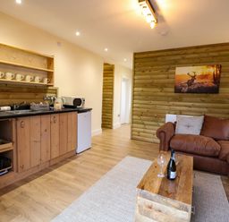 A kitchen and living area with wooden cabinets a brown sofa a wooden coffee table with wine glasses and a bottle on it and a painting of a deer on the wall at Rabbit Warren Lodge in Snettisham