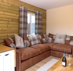A corner sofa with cushions next to a window with curtains and a wooden table with a bottle and glasses at Robins Nest Lodge in Snettisham