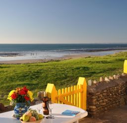 A patio with a table holding flowers fruit and wine glasses overlooking a grassy area and a beach at Homestead Aberdesach near Penygroes