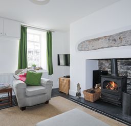 A living room with an armchair next to a window with green curtains and a wood burning stove at Quarrymans cottage in Beddgelert