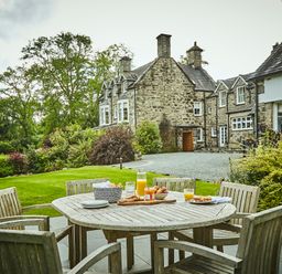 An outdoor patio with a wooden table and chairs set with breakfast items near a stone house surrounded by greenery at Tan Llan in Llanelltyd near Dolgellau