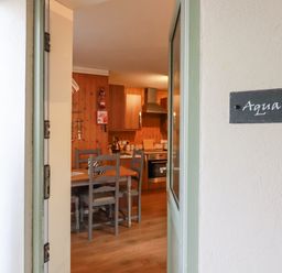 View through an open door into a kitchen dining area with wooden chairs and table and kitchen cabinets at The Aqua Shack in Newquay