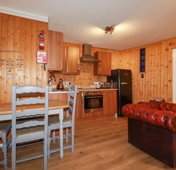 A kitchen and dining area with wooden walls a black fridge wooden cabinets a wooden table with gray chairs and a red leather sofa at The Aqua Shack in Newquay