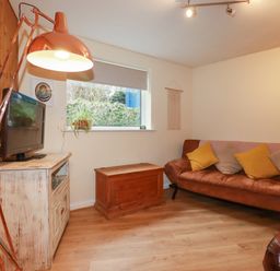 A living room with a brown leather sofa yellow and beige cushions a wooden chest a white distressed cabinet with a television and a large copper lamp at The Aqua Shack in Newquay