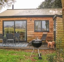 A wooden cottage exterior with a deck table and chairs a barbecue grill and a dog on the grass at Holm Oaks Cottage in Busbridge near Godalming