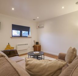 A living room with a beige sofa two cushions a round coffee table a small wooden cabinet a window with a closed blind and a wall-mounted television at Hub House in Three Oaks near Hastings