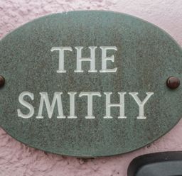 A green oval house name plaque with the text The Smithy on a pink textured wall at The Smithy in Chathill near Seahouses