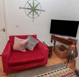 A red armchair with two pillows next to a wooden TV stand with a flat screen TV and a woven basket in a living room at The Smithy in Chathill near Seahouses