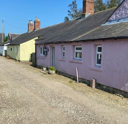 A row of single-story cottages with pink yellow and white walls along a gravel driveway at The Smithy in Chathill near Seahouses