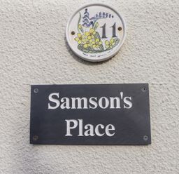 A wall with a house number 11 plaque decorated with flowers and a black sign reading Samsons Place at Morfa Bychan