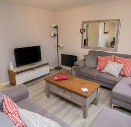 A living room with a gray sectional sofa with patterned cushions a wooden coffee table a television on a stand a floor lamp and a large mirror on the wall at Lewis Lodge in St Ann's Chapel