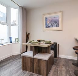 A dining area with a wooden table set for four and cushioned stools near a window with curtains at Hollycombe in Ilfracombe