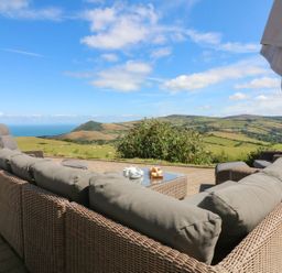 An outdoor seating area with wicker furniture and cushions overlooking fields and hills at Buddicombe House in Combe Martin