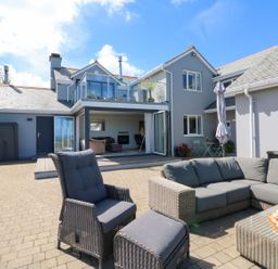 An outdoor patio area with gray cushioned wicker furniture and a modern house with a balcony at Buddicombe House in Combe Martin
