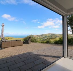 An outdoor patio with wicker furniture and umbrella overlooking green hills and sea at Buddicombe House Combe Martin
