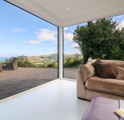 A corner of a living room with a sofa and glass walls showing an outdoor patio and countryside view at Buddicombe House in Combe Martin