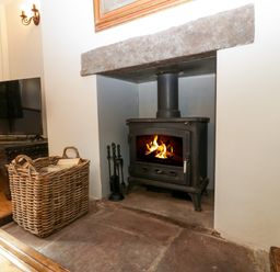 A room with a wood burning stove in a stone hearth with a basket of firewood and a television on a wooden table at Court House Cottage in Gatcombe near Blakeney