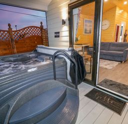 An outdoor hot tub next to a sliding glass door leading to a wooden interior with a sofa and a bed at Binevanagh Cabin in Magilligan near Castlerock
