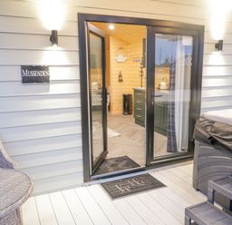 An open glass door leading to a kitchenette with cabinets and a kettle outside on a wooden deck with a wicker chair table and a hot tub at MUSSENDEN CABIN in Magilligan near Castlerock