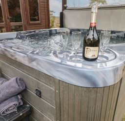 An outdoor hot tub with a bottle of prosecco and three glasses on the edge near rolled towels at Greenhaven in Narberth