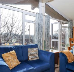A living room with blue sofas and patterned cushions next to large windows and glass doors at Matilda Lodge in Newton Ferrers