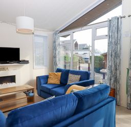 A living room with blue sofas a wooden coffee table a wall-mounted television and large windows with curtains at Matilda Lodge in Newton Ferrers