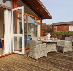 A patio with wicker table and chairs set with drinks and pastries outside a house with glass doors at Matilda Lodge in Newton Ferrers