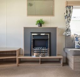 A living room with a fireplace a wooden shelf a plant a window with curtains and a gray sofa with patterned cushions at Ripples & Reeds - Billing Aquadrome