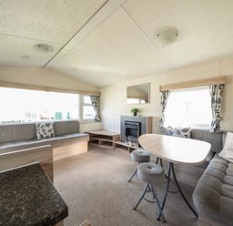 A living area with two sofas a wooden table two stools a fireplace and windows with curtains at Ripples & Reeds - Billing Aquadrome in Billing Aquadrome Holiday Park