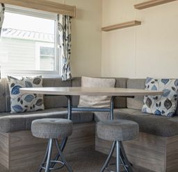 A dining area with a wooden table surrounded by gray cushioned bench seating and two stools at Ripples & Reeds - Billing Aquadrome in Billing Aquadrome Holiday Park