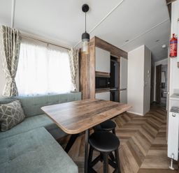 A dining area with a wooden table and two black stools beside an L-shaped sofa with cushions and a window with curtains at Waterslide Way - Billing Aquadrome in Billing Aquadrome Holiday Park