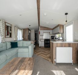 A living area with a light green sofa and wooden coffee table next to a kitchen with wooden cabinets and appliances at Stillwater Retreat - Billing Aquadrome Holiday Park