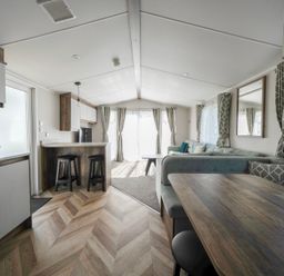 An open plan living space with a kitchen area including stools and a toaster a dining area with a wooden table and cushioned seating and a seating area with sofas and curtains at Stillwater Retreat Billing Aquadrome Holiday Park