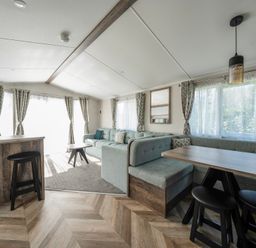 A living room with a dining area a sofa a coffee table bar stools and windows with curtains at Stillwater Retreat - Billing Aquadrome in Billing Aquadrome Holiday Park