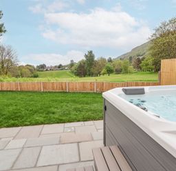 A backyard with a hot tub on a stone patio and a grassy lawn enclosed by a wooden fence near a hill at Ty Blodau near Prestatyn