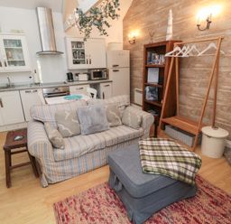 A small living area with a striped sofa, ottoman with a blanket, wooden bookcase, clothing rack, and kitchen in the background at The Forge in Nr Seashouses