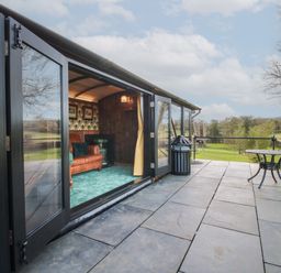 An outdoor patio with stone tiles and a small round table next to a glass door leading to a room with a leather sofa and green carpet at GWR Wagon - Victoria Station in Ludlow