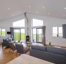 A living room with gray sofas chairs a wood burning stove and large windows at Jacana in Perranporth