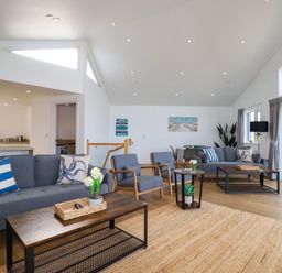 A living room with two blue sofas, two blue armchairs, coffee tables, and a kitchen area at Jacana Perranporth