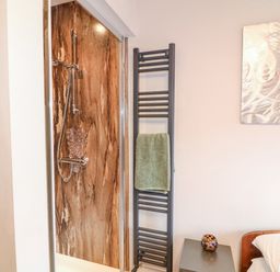A bathroom shower with a brown marble wall panel next to a black towel radiator with a green towel and part of a bedroom with a bedside table and an artwork on the wall at Orchard Retreat Staycation in Kirton