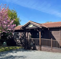 A wooden house with a covered entrance and tiled roof next to a pink flowering tree at Orchard Retreat Staycation in Kirton