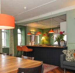 A dining area with wooden tables and chairs a bar with orange stools and orange pendant lights and a grey sofa with cushions at Sycamore Apartment in Lifton