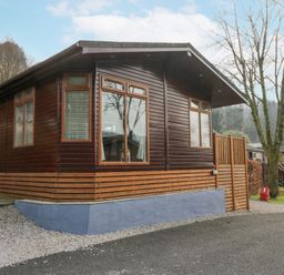 A wooden cabin with multiple windows next to a gravel path and trees at Swifts Reach Lodge Kirkstone 10 Limefitt Park near Windermere