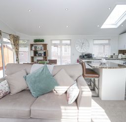 A living room with a sofa and cushions next to a kitchen island with chairs and a wall clock at Swifts Reach Lodge Kirkstone 10 Limefitt Park near Windermere