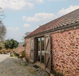 An exterior view of a stone building with wooden doors open on a gravel ground with trees in the background at The Barn in Spaxton
