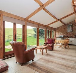 A living area with two leather armchairs a wooden coffee table a dining table with chairs kitchen cabinets and large windows showing greenery outside at The Barn in Spaxton