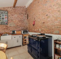 A kitchen with a stone wall, sink, microwave, stove, wooden chairs and shelves with cups and glasses at The Barn in Spaxton