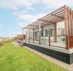 A wooden deck with a pergola and outdoor dining furniture next to a grassy area at Hepworth in Portreath