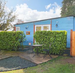 A small patio with a table and two chairs outside a blue wooden building with green hedges at Shed Your Blues Brilley near Kington